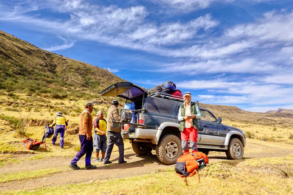 Group of people preparing for an outdoor adventure, with backpacks and gear around a parked SUV in a mountainous landscape.