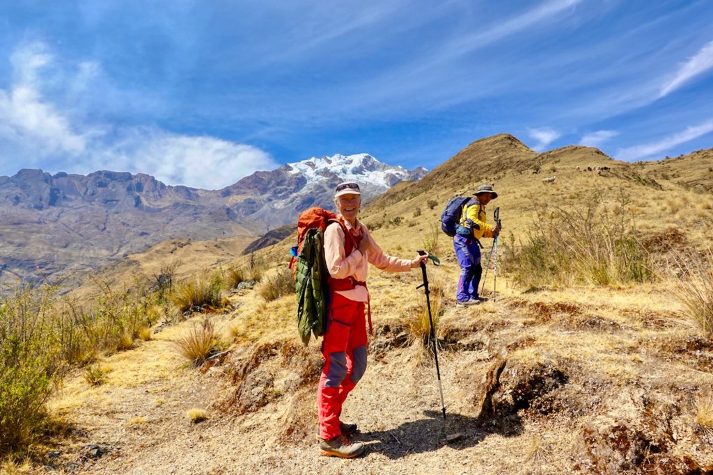 Two hikers on a mountainous trail with snow-capped peaks in the background, one smiling and holding trekking poles.