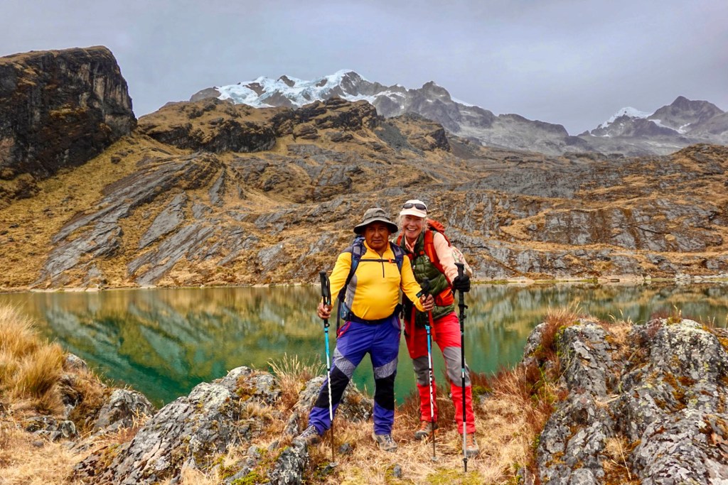 Two hikers posing together by a reflective mountain lake, surrounded by rocky terrain and snow-capped peaks.