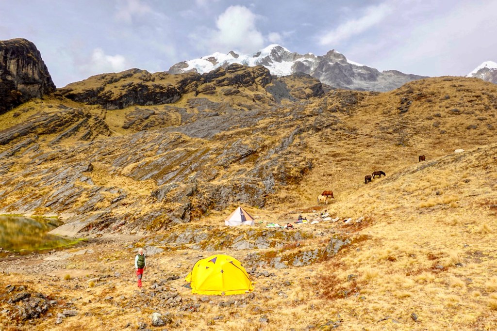 A scenic camping site in the mountains with a yellow tent and a pink tent, surrounded by rocky terrain and horses grazing in the distance. Snow-capped peaks are visible in the background.
