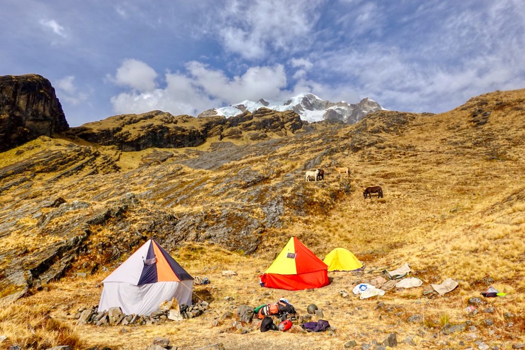 Camping tents set up on rocky terrain with a mountainous backdrop and grazing horses in a grassy field.