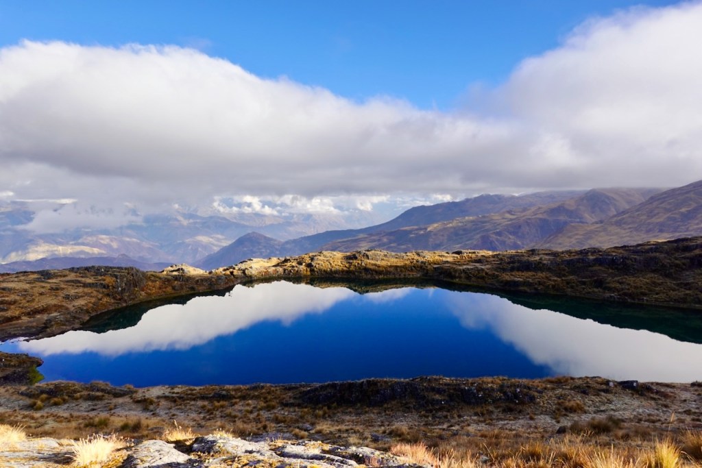 A serene mountain lake reflecting the sky and surrounding hills, with rocky terrain and patches of grass.