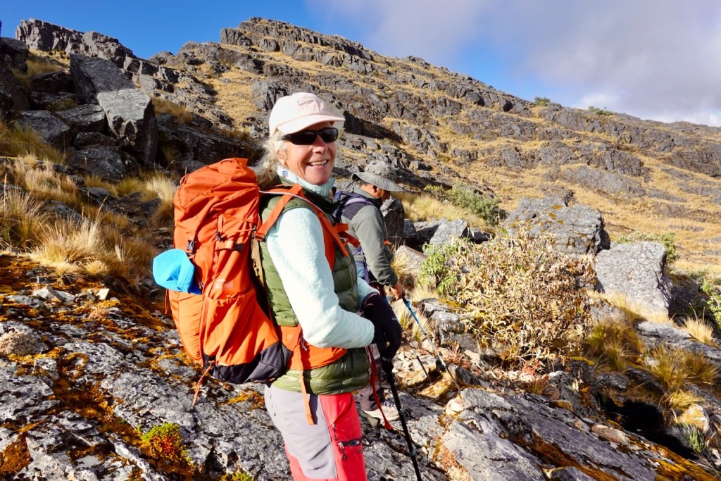 A smiling woman wearing a hat and sunglasses stands on rocky terrain, carrying an orange backpack, with a male hiker in the background.