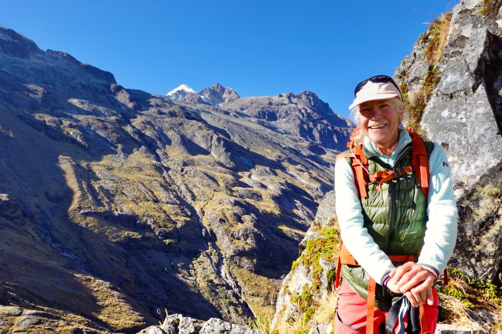 A smiling woman wearing hiking gear stands on a rocky mountain trail, with a backdrop of steep, rugged mountains and a blue sky.