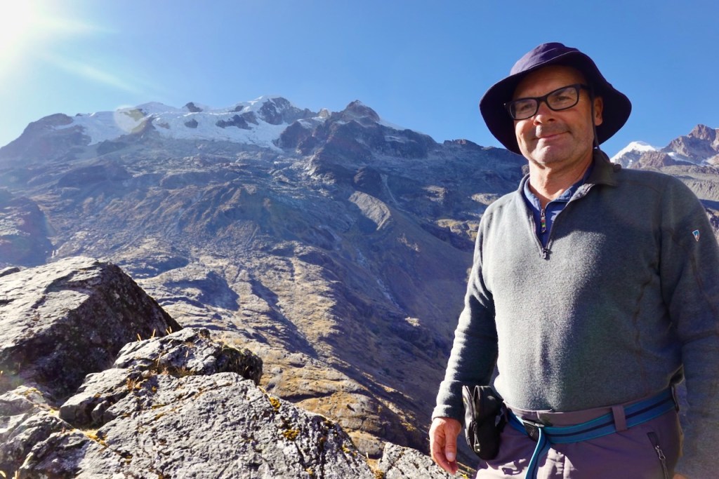 A man wearing a hat and glasses stands on a rocky terrain with a mountainous backdrop, including snow-capped peaks under a clear blue sky.