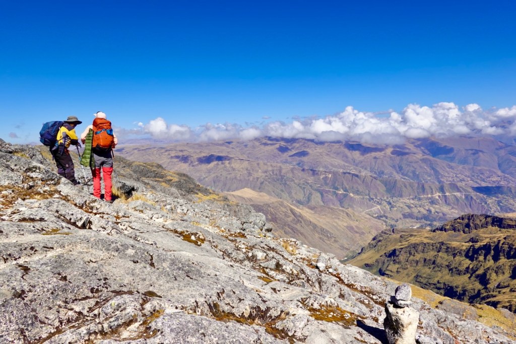 Two hikers navigating rocky terrain on a mountain peak, overlooking a vast landscape with valleys and distant mountains under a clear blue sky.