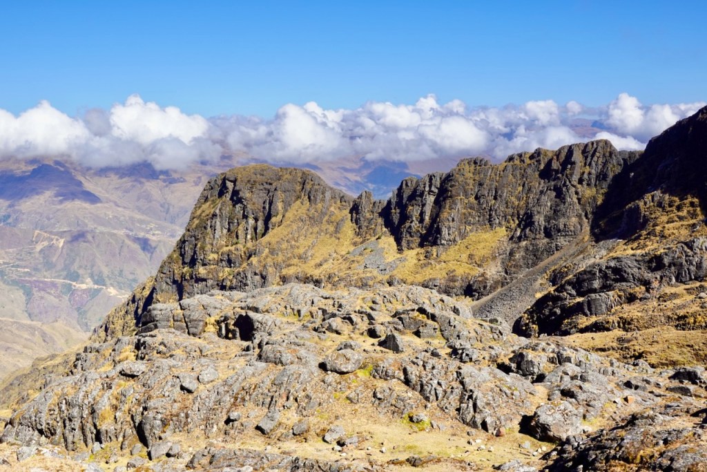 Panoramic view of rocky mountain terrain under a clear blue sky, with scattered clouds in the background.