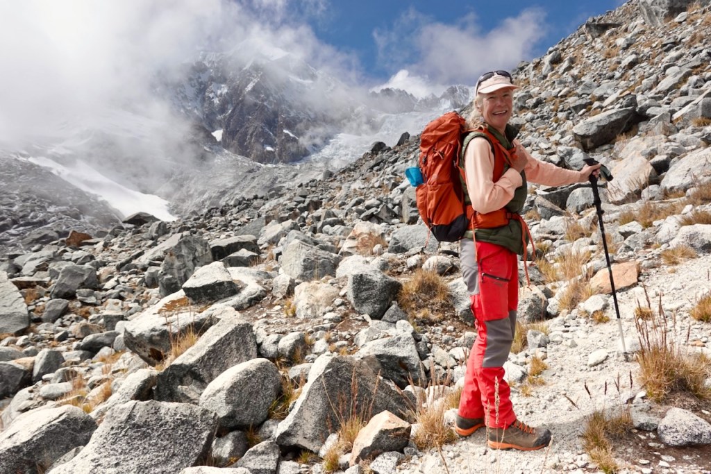A hiker standing on a rocky mountain trail, wearing a backpack and using a walking stick, with a mountainous backdrop covered in clouds.