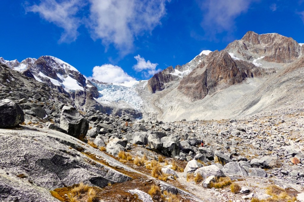 A rugged mountainous landscape featuring rocky terrain, glaciers, and a clear blue sky with scattered clouds.