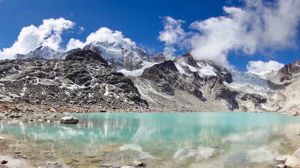 A panoramic view of a turquoise glacial lake surrounded by rocky mountains and snow-capped peaks under a bright blue sky with fluffy white clouds.