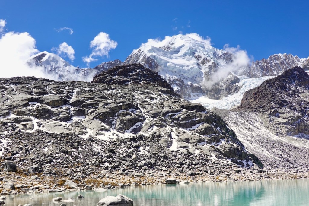 A scenic view of a mountainous landscape featuring snow-capped peaks, rocky terrain, and a clear turquoise lake under a bright blue sky with scattered clouds.
