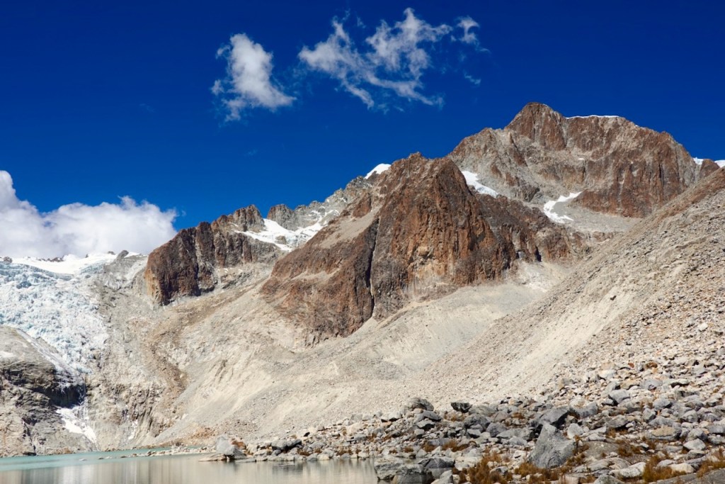 A panoramic view of rugged mountain peaks against a bright blue sky, featuring rocky terrain and patches of snow.