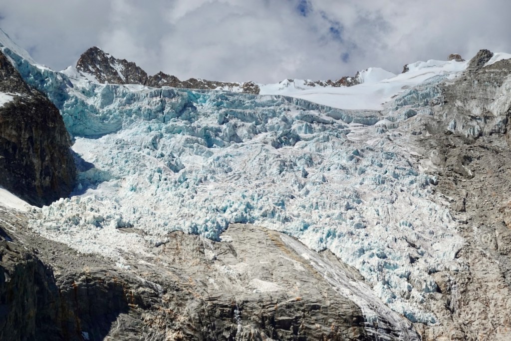 A close-up view of a glacier cascading down a rocky mountain, featuring bright blue ice and rugged terrain under a cloudy sky.