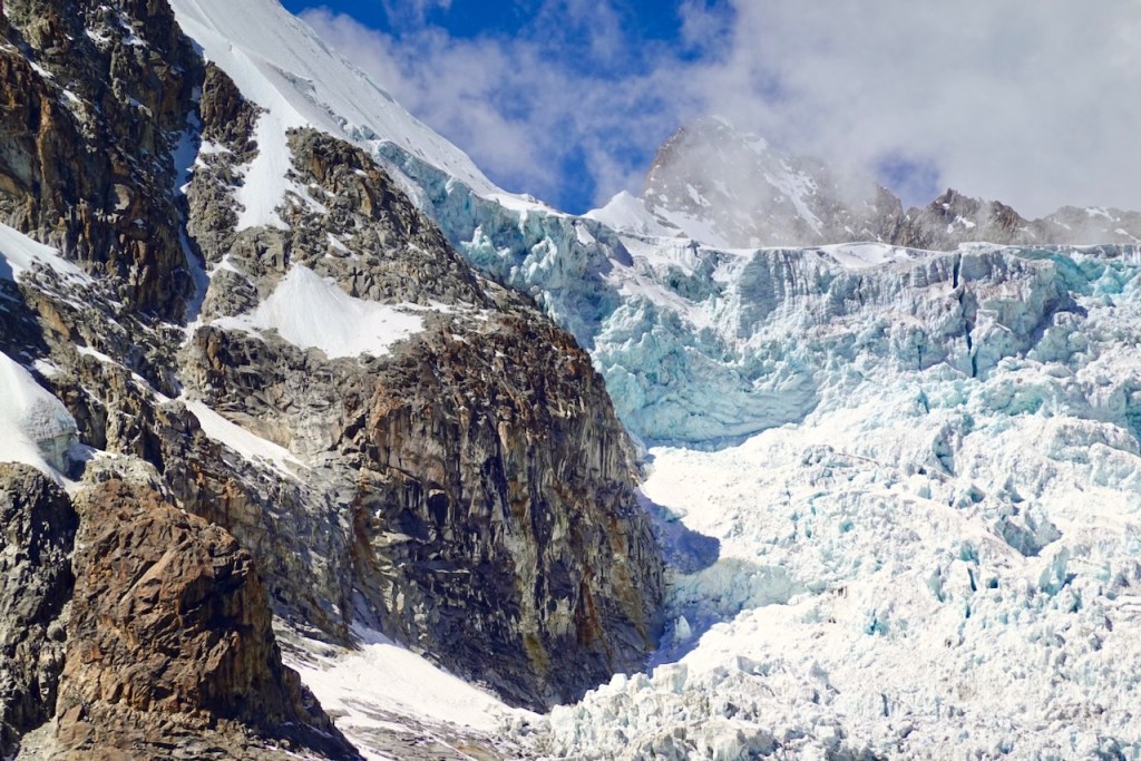 A close-up view of a glacier on a rocky mountain, showcasing icy blue and white ice formations amidst steep, rugged terrain under a partly cloudy sky.