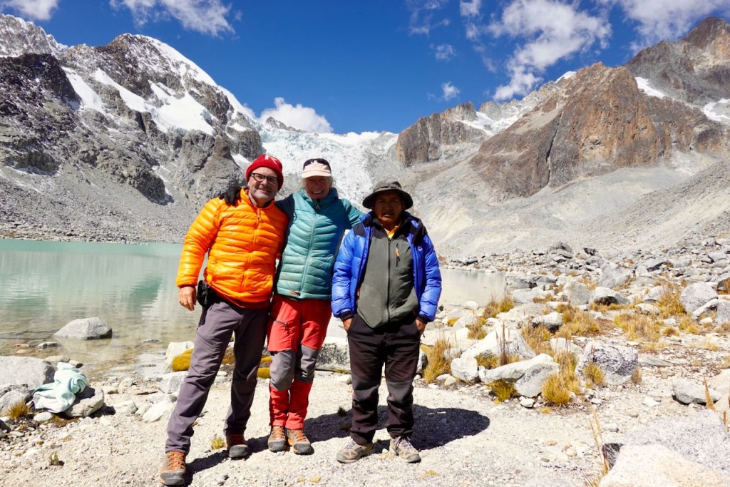 Three hikers posing together near a glacial lake with mountains in the background.