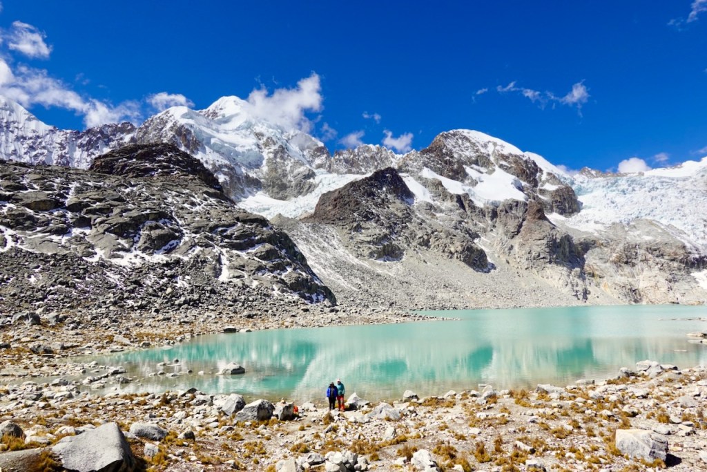 Two hikers standing near a turquoise glacial lake surrounded by rocky mountains and snow-capped peaks under a bright blue sky.