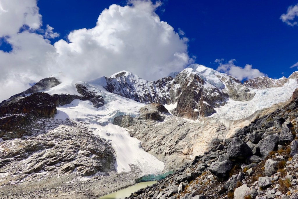 A panoramic view of rugged snow-capped mountains under a bright blue sky with fluffy clouds, showcasing rocky terrain and a glacial lake in the foreground.