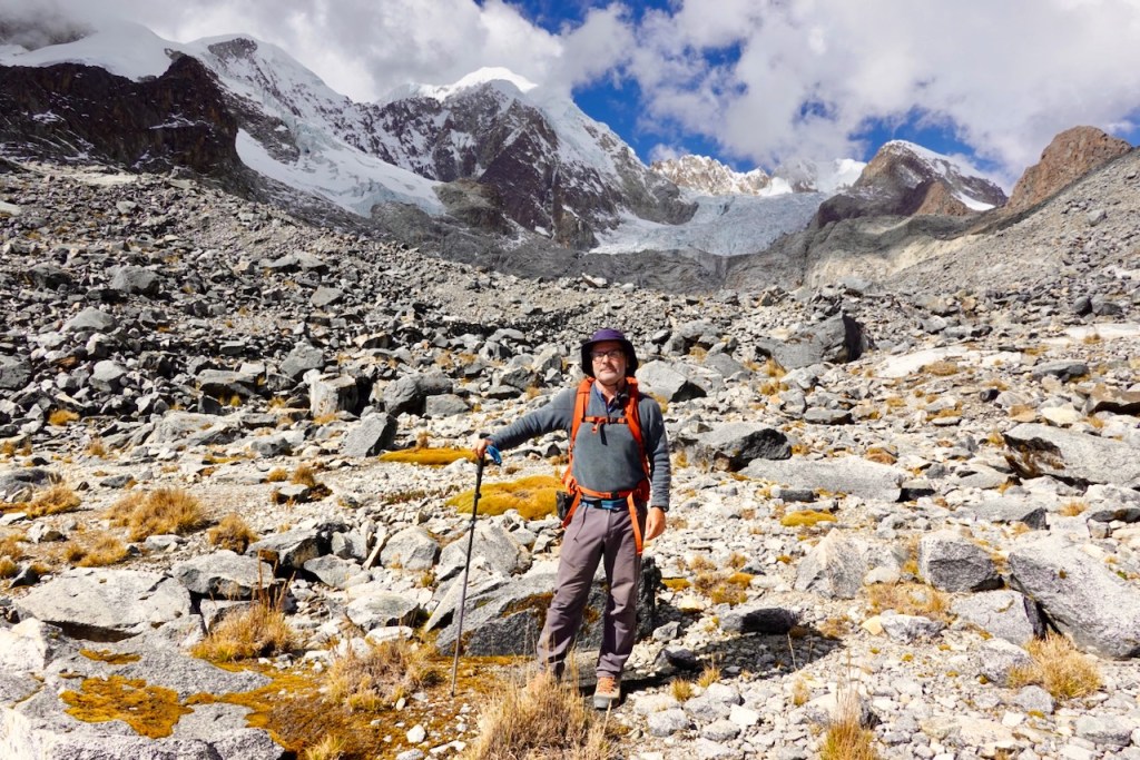 A hiker standing on rocky terrain with mountains and a glacier in the background, dressed in outdoor gear and holding a trekking pole.