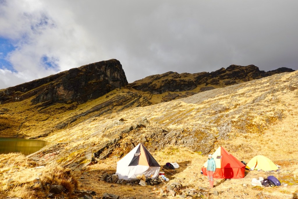 Campsite with colorful tents set against a mountainous landscape and a lake, under a partly cloudy sky.