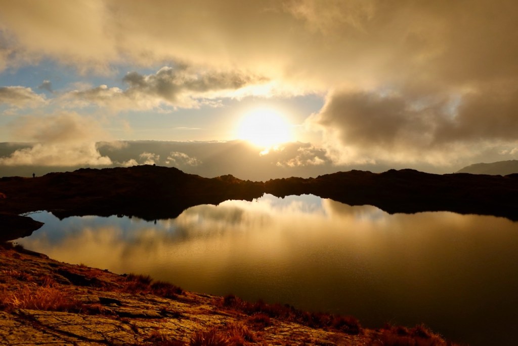 A serene landscape featuring a sunset over a still lake, with clouds reflecting in the water, and rocky terrain visible in the foreground.