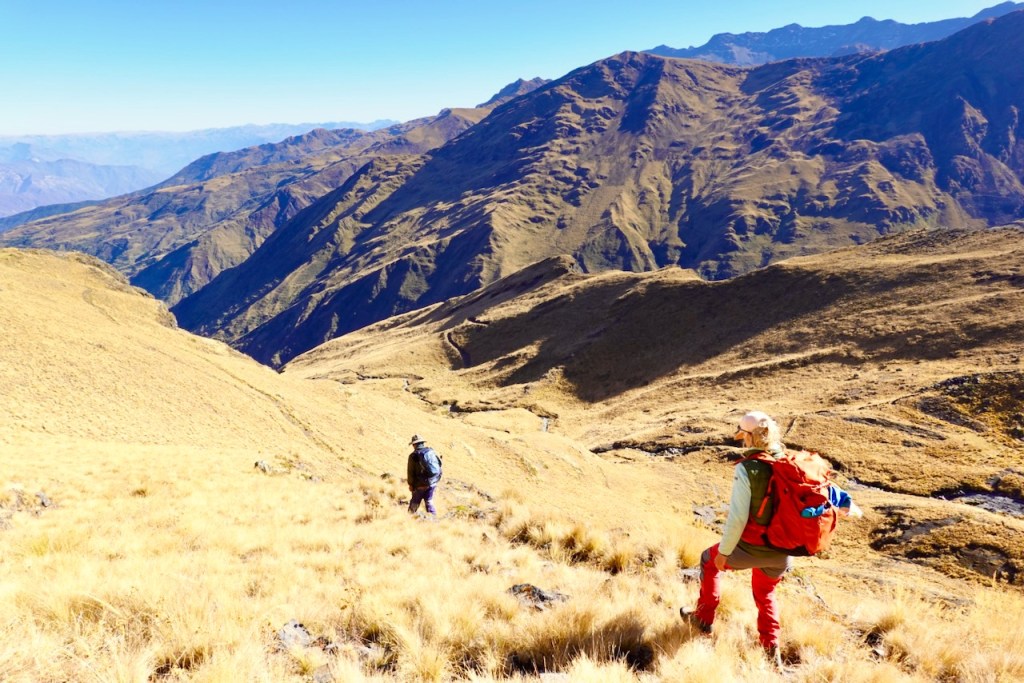 Two hikers ascending a grassy hillside with mountains in the background under a clear blue sky.