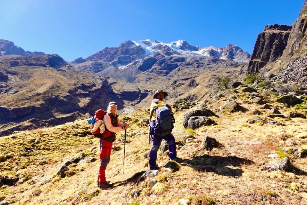 Two hikers standing on a grassy hillside with rocky terrain and snow-capped mountains in the background under a clear blue sky.