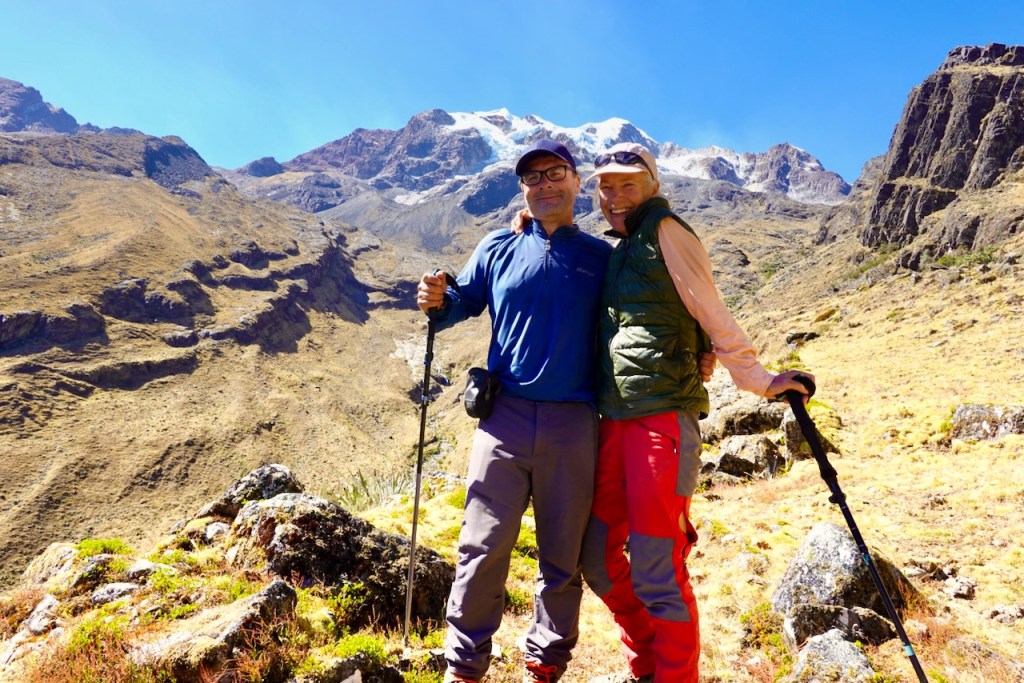 A couple stands together smiling on a hiking trail, with mountains and rugged terrain in the background. They are dressed in outdoor gear, holding trekking poles.