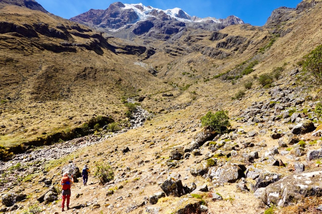 Two hikers trekking through a rocky valley with scattered vegetation, surrounded by steep mountain slopes and a snow-capped peak in the background under a clear blue sky.
