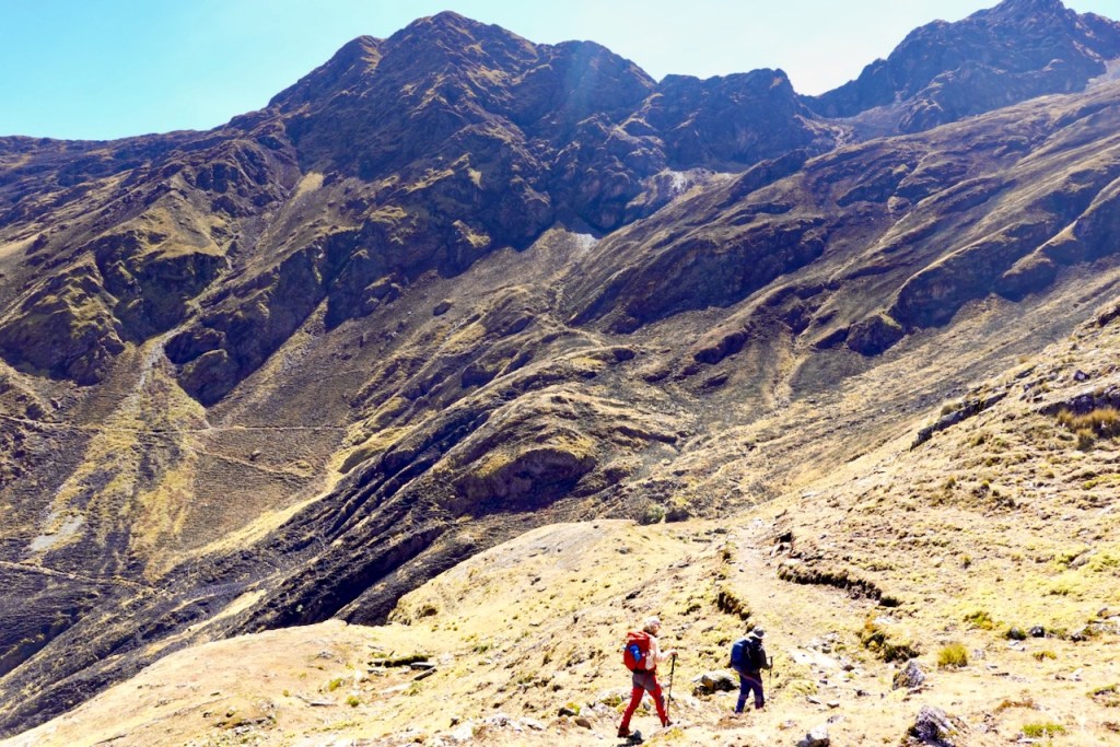 Two hikers trekking on a rocky mountain path surrounded by steep hills and clear blue skies.