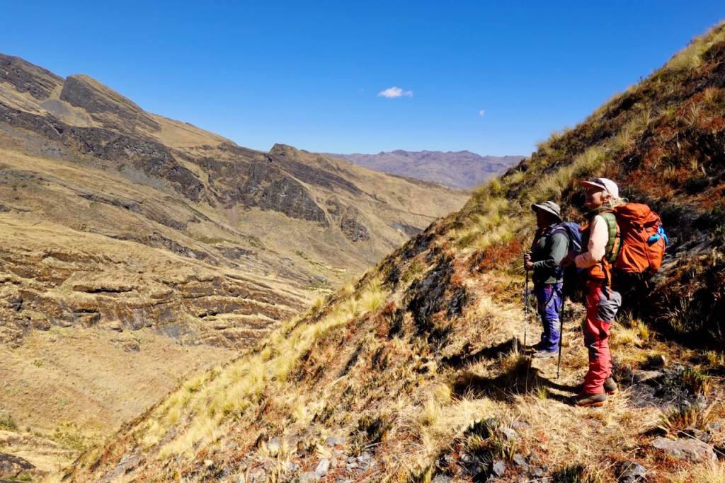 Two hikers standing on a rocky hillside, looking out over a vast mountainous landscape under a clear blue sky.
