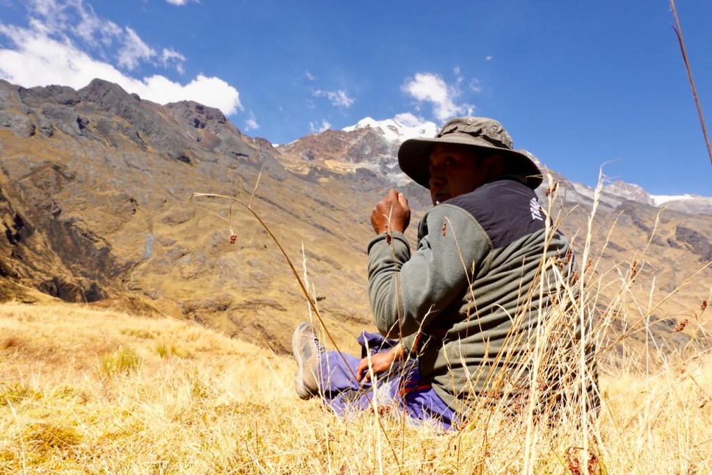 A man sitting on the grass in a mountainous landscape, wearing a wide-brimmed hat and a green jacket, with snow-capped peaks in the background and a clear blue sky.