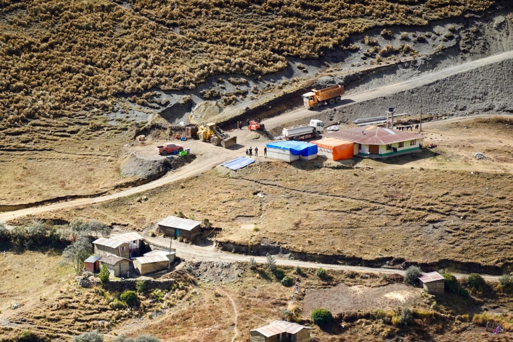 Aerial view of a rural area featuring several buildings, vehicles, and construction equipment on a dirt road surrounded by dry land and sparse vegetation.