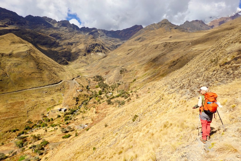 A hiker with a backpack stands on a rocky trail overlooking a valley surrounded by mountains, showcasing a scenic view of the landscape with dry grass and scattered vegetation.