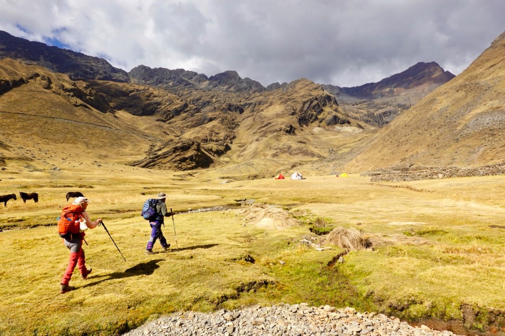 Two hikers walking on a grassy plain with mountains in the background, surrounded by tents and horses.