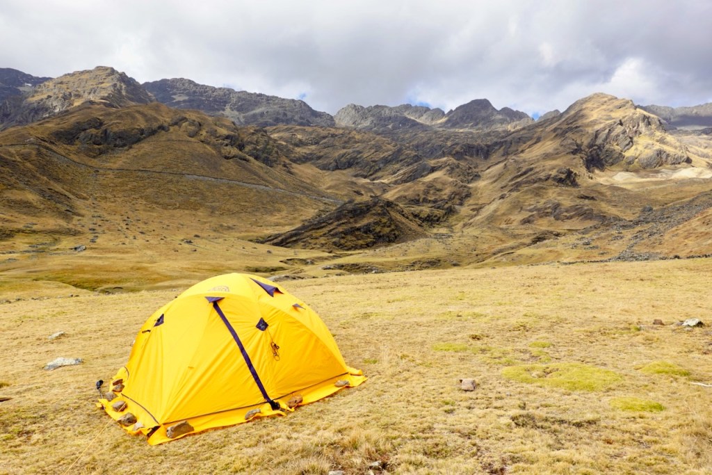 A bright yellow camping tent set against a rugged mountain landscape with rolling hills and cloudy skies.