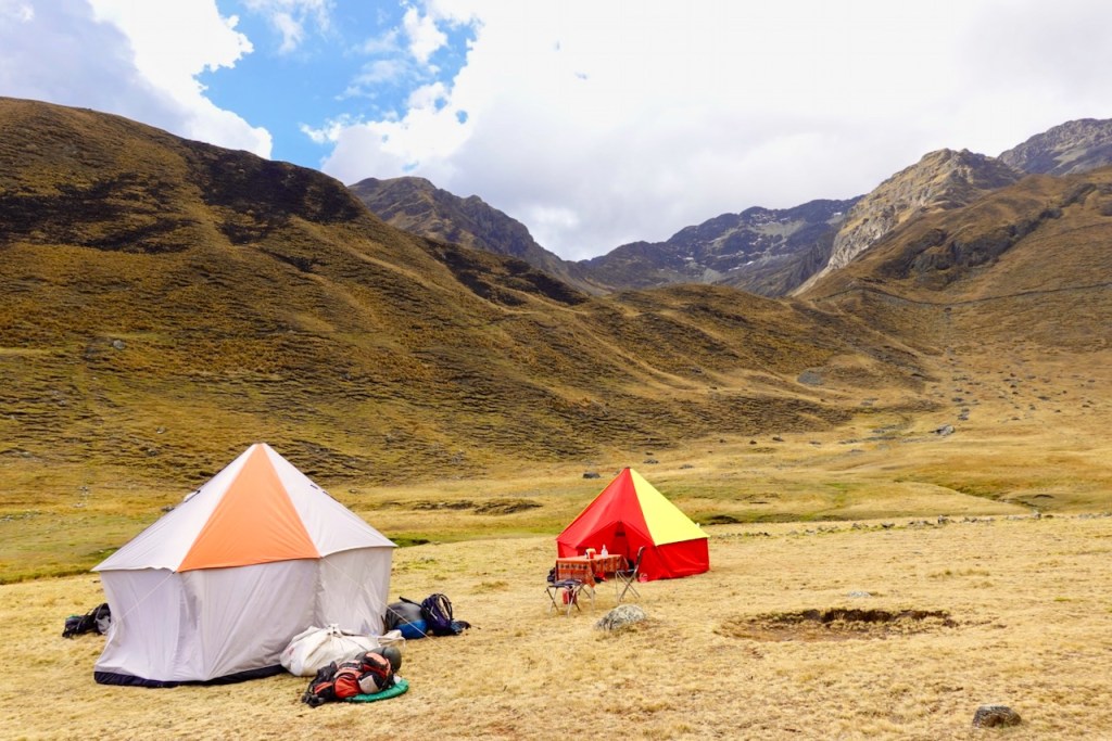 Two tents set up in a grassy area surrounded by mountains under a partly cloudy sky.