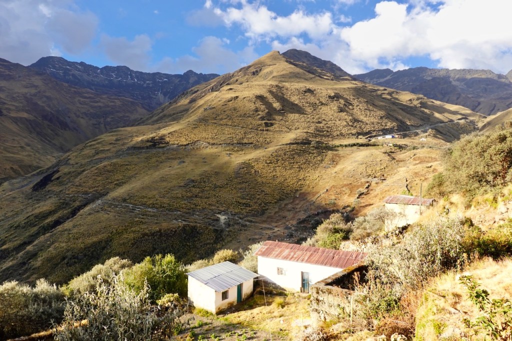 A scenic view of a mountainous landscape featuring two small houses, with dry, rolling hills and rocky peaks under a blue sky with scattered clouds.