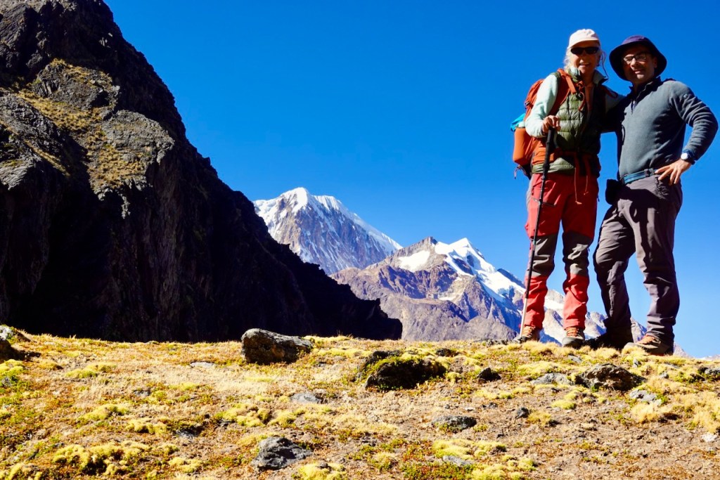 Two hikers standing on a grassy terrain with mountains in the background, wearing hiking gear and smiling.