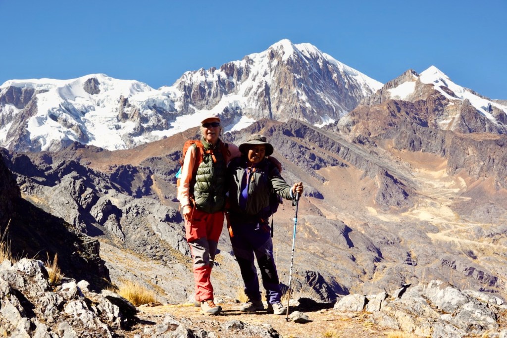Two hikers posing on a rocky mountain trail with snow-capped peaks in the background.