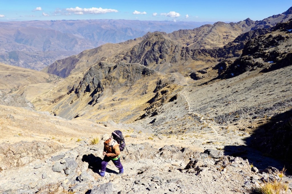 A hiker climbing a rocky mountain trail with expansive valleys and mountains in the background under a clear blue sky.