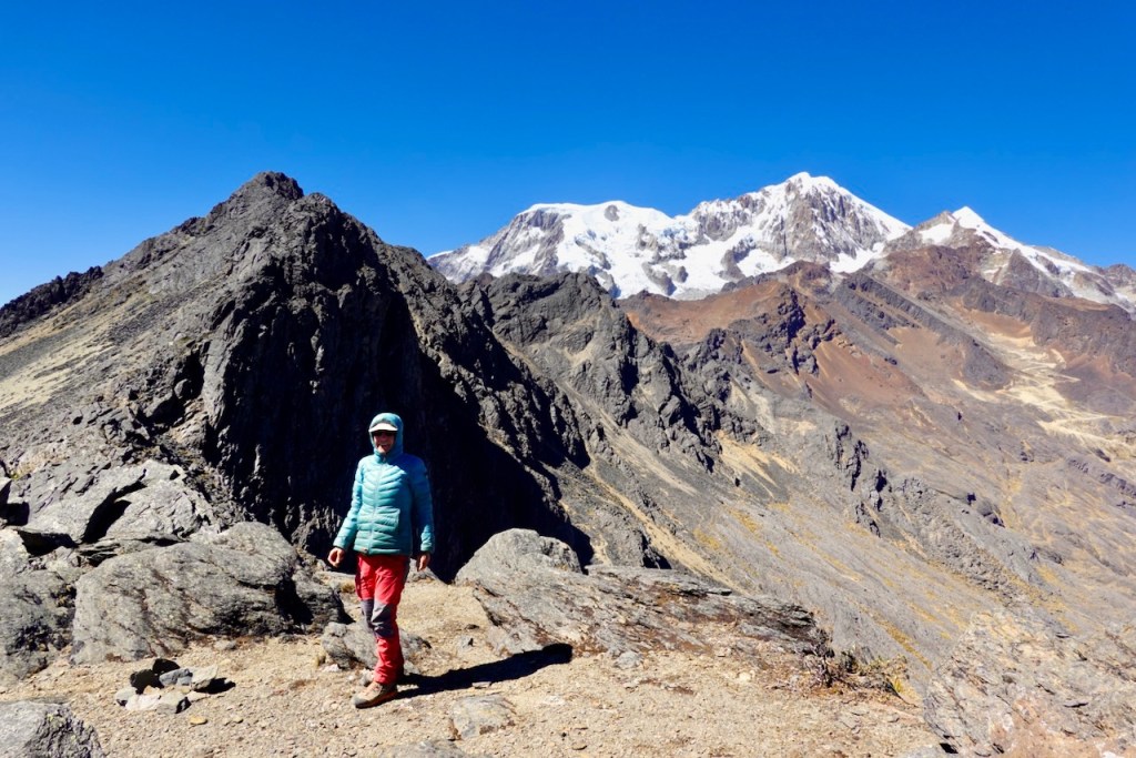 A person in a blue jacket and red pants stands on a rocky mountain edge with a snow-capped peak in the background under a clear blue sky.