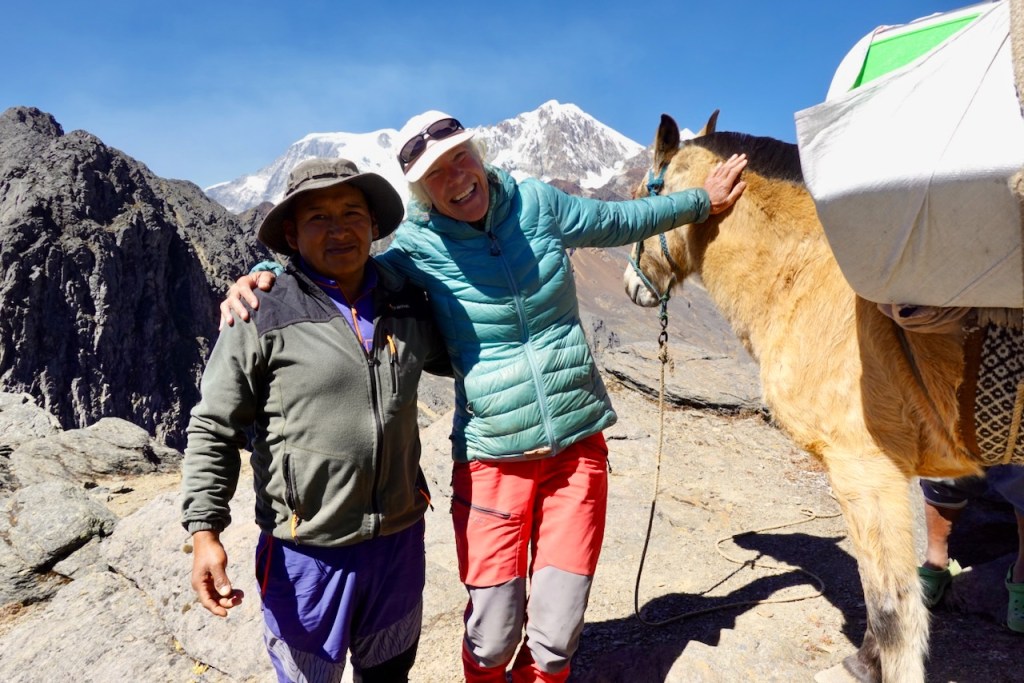 A smiling man and woman pose together in a mountainous landscape, with snow-capped peaks in the background. The woman is wearing a blue jacket, and the man is in a gray sweater. A horse is nearby.