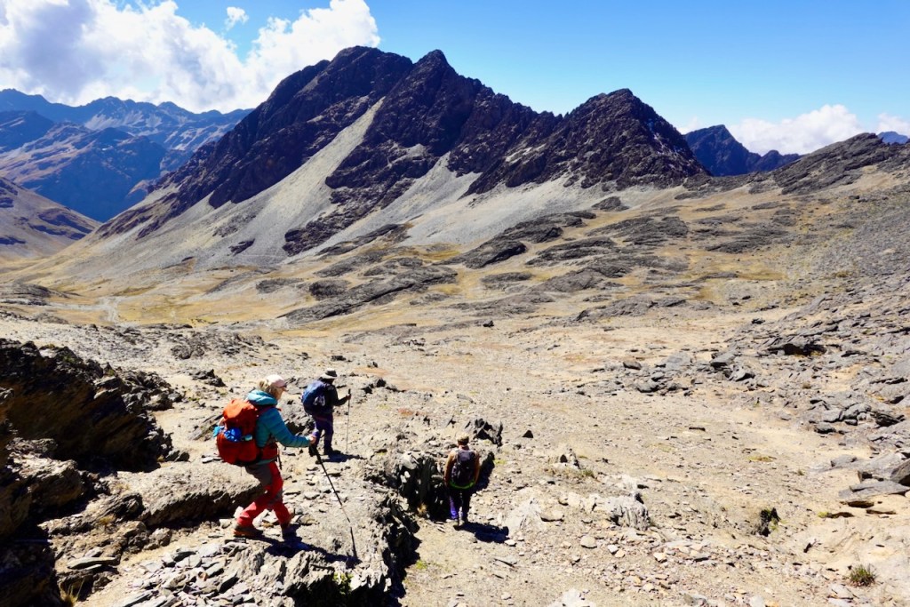 Three hikers navigating rocky terrain in a mountainous landscape under a clear blue sky.