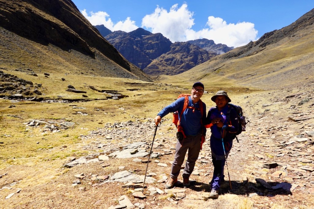 Two hikers smiling while standing on a rocky trail in a mountainous landscape with blue skies and scattered clouds.
