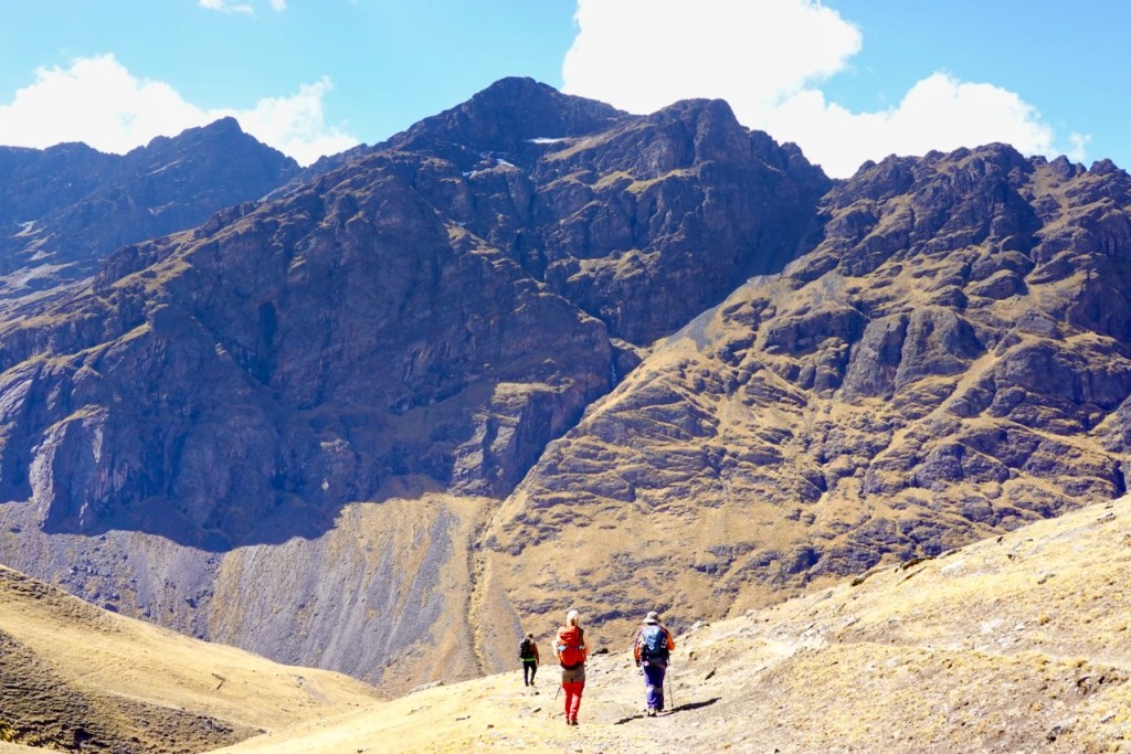 Three hikers walking on a mountain trail with rugged peaks in the background under a blue sky.