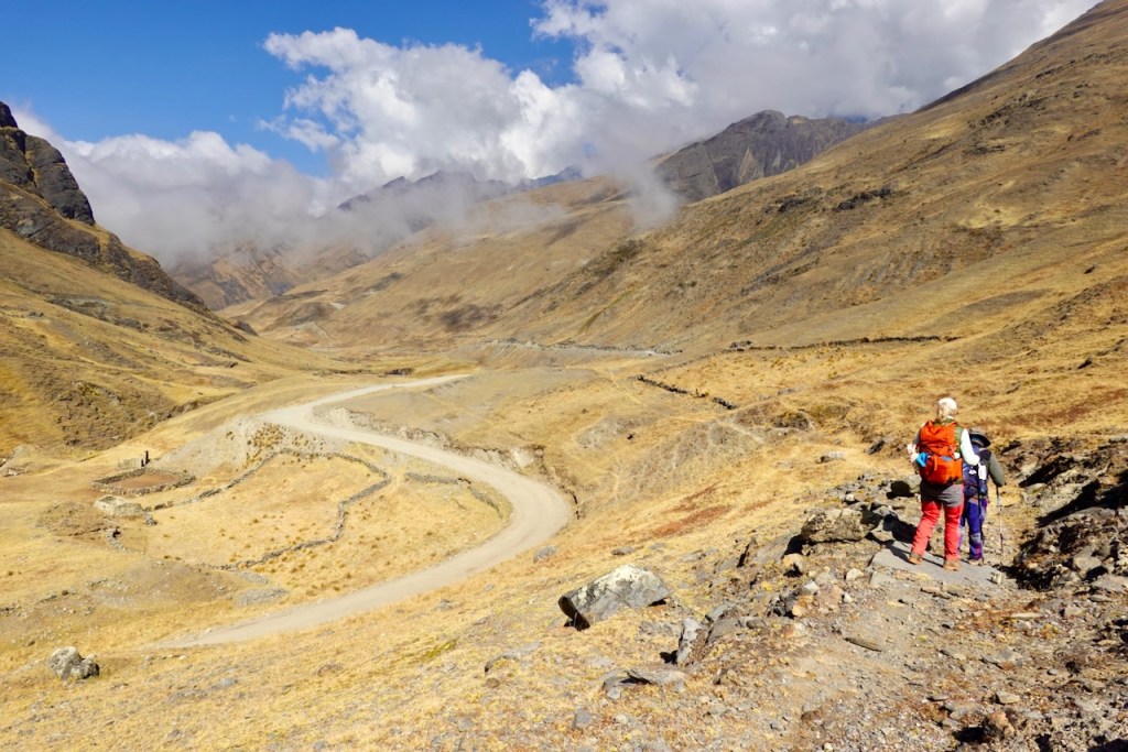 Two hikers standing on a rocky trail overlooking a winding dirt road in a mountainous area with dry grass and scattered clouds in the background.