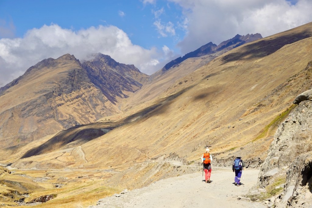 Two hikers walking on a dirt path through a mountain landscape with rocky hills and a blue sky.