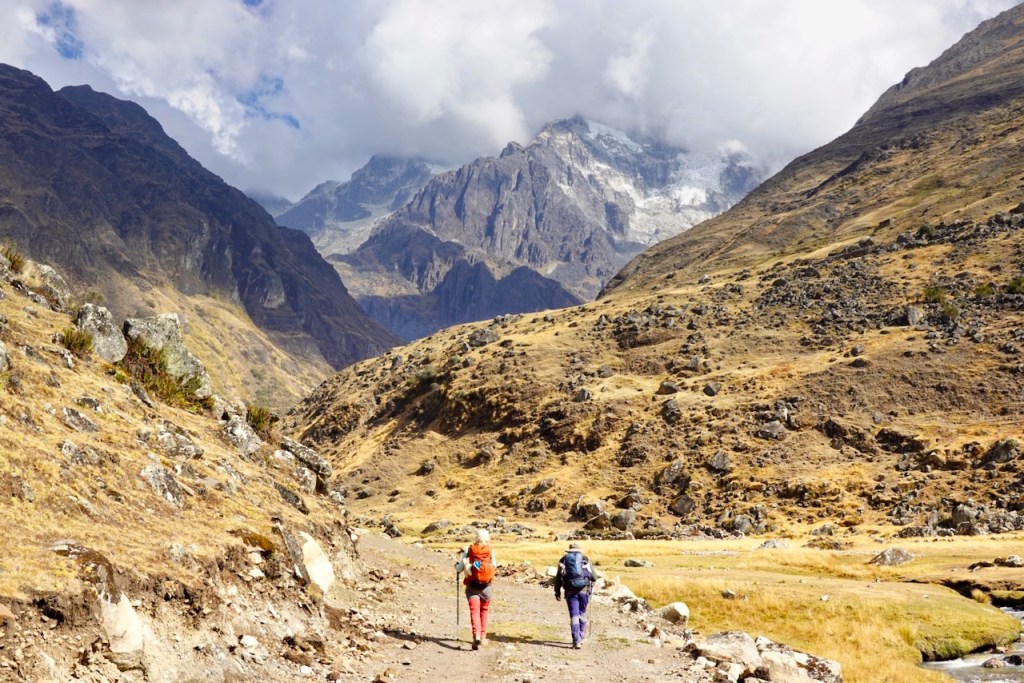 Two hikers walking along a rocky trail in a mountainous landscape, surrounded by steep cliffs and snow-capped peaks under a partly cloudy sky.