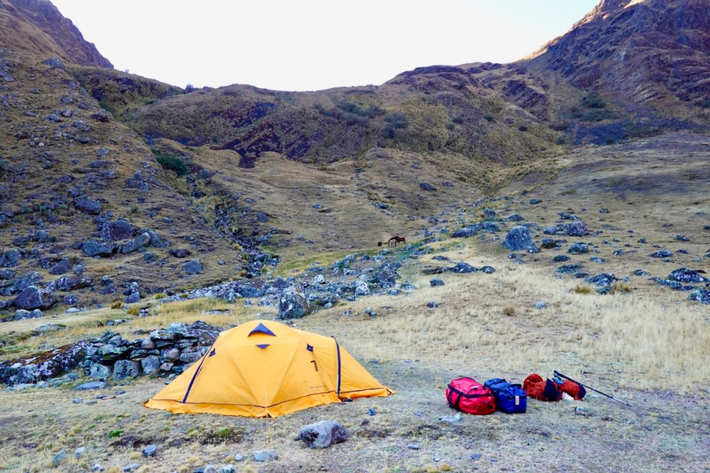 A yellow camping tent set up in a mountainous landscape with brown grass and scattered rocks. Several backpacks are placed beside the tent.