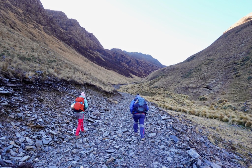 Two hikers walking on a rocky trail in a mountainous area, surrounded by steep slopes and grass-covered terrain.